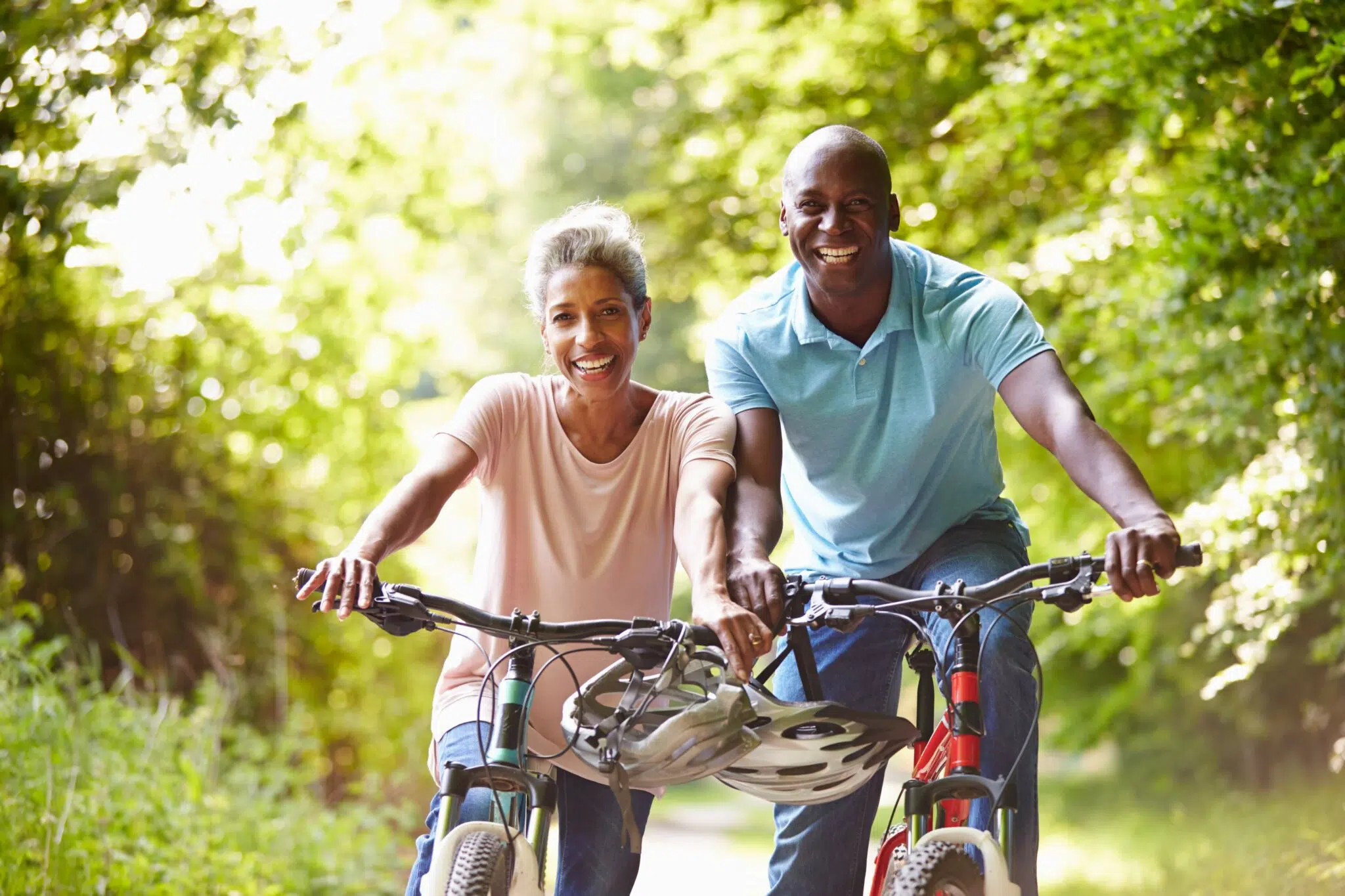 Two adults riding bicycles outdoors on a sunny day, smiling and enjoying a green, tree-lined path, with a helmet resting on each of their bike's handlebars.